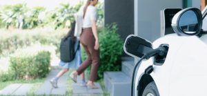 Electric vehicle recharging at home charging station plugged in with EV charger device with blurred background of progressive mother and daughter walking as concept for sustainability of energy.