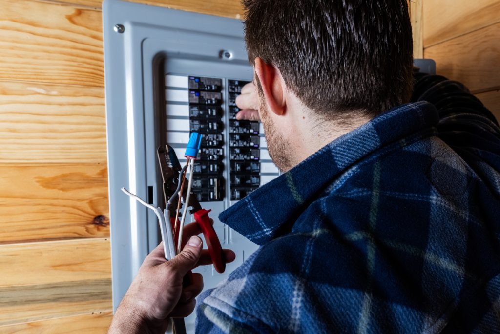 An over-the-shoulder image of a man repairing a gray electrical breaker box.