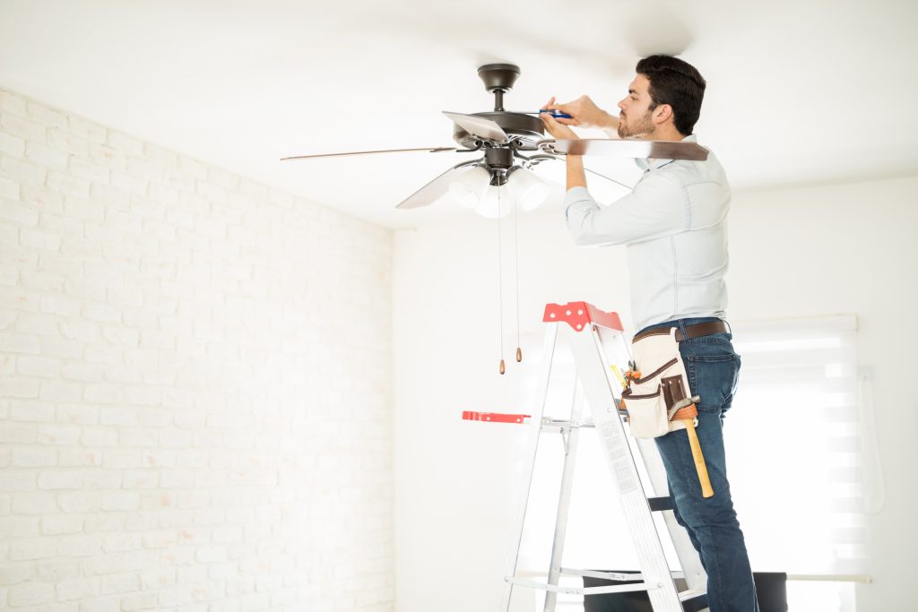 A professional electrician installs a ceiling fan in a bright sunny room. 