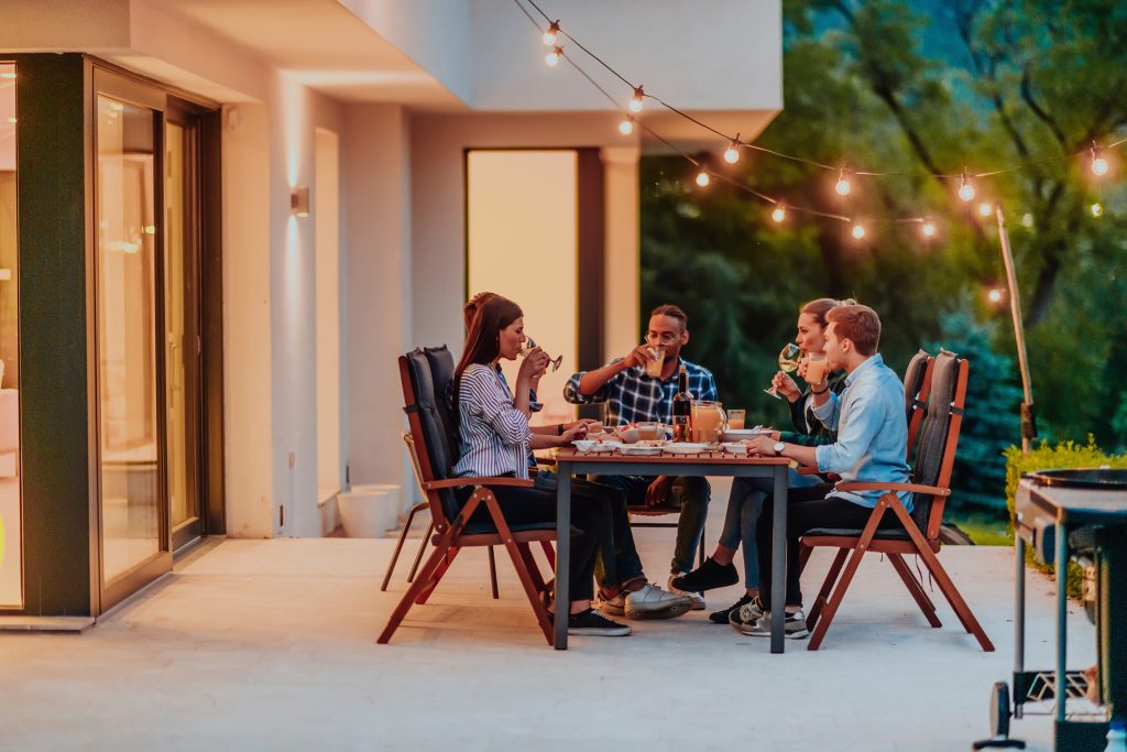 Friend group having dinner on a well-lit patio outside a beautiful home with energy-efficient lighting indoors.