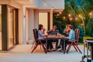 Friend group having dinner on a well-lit patio outside a beautiful home with energy-efficient lighting indoors.